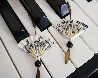 Könnte beinhalten: Ein Paar Ohrringe mit einem Musiknoten-Fächerdesign. Die Fächer bestehen aus weißem Papier mit schwarzen Noten darauf gedruckt. Die Ohrringe haben schwarze Perlen und bronzefarbene Akzente.