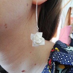 May include: A pair of silver dangle earrings with a white origami flower charm. The earrings are worn by a person with a blue and white patterned shirt.