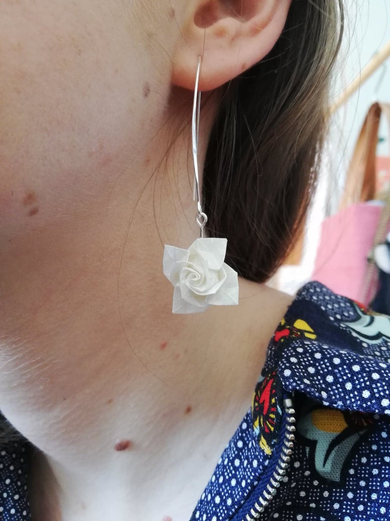 May include: A pair of silver dangle earrings with a white origami flower charm. The earrings are worn by a person with a blue and white patterned shirt.