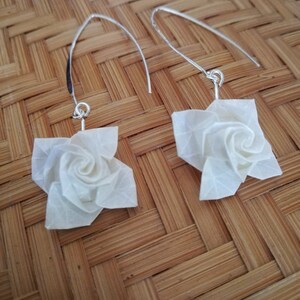 May include: A pair of silver earrings with white origami rose flower charms. The earrings are hanging from a woven bamboo surface.