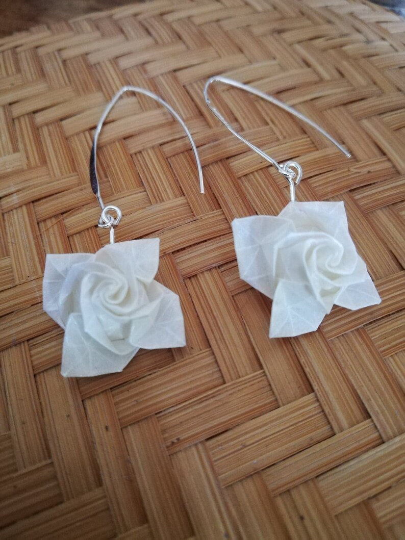 May include: A pair of silver earrings with white origami rose flower charms. The earrings are hanging from a woven bamboo surface.