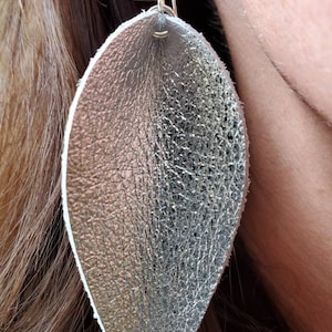 May include: Silver leaf-shaped earrings with a textured, metallic finish. The earrings are suspended from a silver hook. The design is a teardrop shape, with a white edge. The earrings are lightweight and suitable for everyday wear.