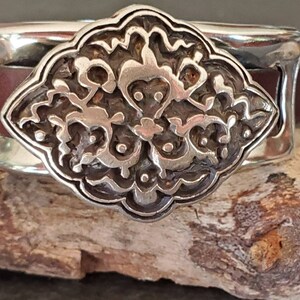 May include: A silver cuff bracelet with a decorative, ornate, silver design in the center. The bracelet is made of metal and has a brown leather band.