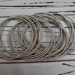 May include: A collection of silver-colored metal bangles, stacked and arranged in a circular pattern. The bangles are thin and have a textured surface. The background is a white, weathered wooden surface.
