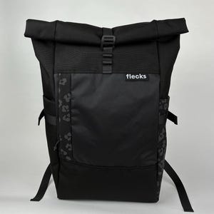 Könnte beinhalten: Schwarzer Rucksack mit Rollverschluss, einer Vordertasche und Seitentaschen. Der Rucksack hat ein Leopardenmuster auf der Seite und das Wort "flecks" auf der Vordertasche. Verstellbare Träger und ein Schnallenverschluss sind sichtbar.