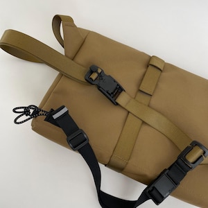 Op de afbeelding: Een bruine messenger bag met zwarte en olijfgroene riemen en gespen. De tas heeft een rechthoekige vorm met een klepsluiting en verstelbare riemen. De tas is gemaakt van een duurzaam, waterbestendig materiaal.