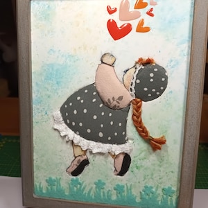 Peut inclure: Une œuvre d'art encadrée représentant une fillette en tissu, vêtue d'une robe grise à pois blancs et d'une longue tresse. Le fond est texturé, vert et bleu, avec des cœurs. Le cadre est gris.