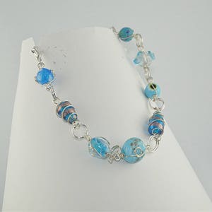 Puede incluir: Un brazalete de cadena de plata con cuentas de vidrio azul y blanco. Las cuentas est&aacute;n envueltas en alambre de plata y tienen una variedad de patrones.