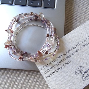 Peut inclure: Un bracelet wrap à plusieurs brins avec un mélange de perles violettes, transparentes et argentées, et de petits accents de pierres irrégulières. Le bracelet est enroulé sur un ordinateur portable. Une carte avec du texte français et un logo est à côté.