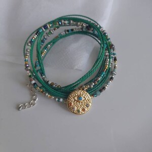 May include: A green and silver beaded bracelet with a gold evil eye charm. The bracelet is made of multiple strands of thread and beads.