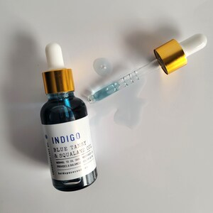May include: A dark blue glass bottle of Indigo Blue Tansy & Squalane Oil with a white label and gold cap. A glass dropper with a gold top is next to the bottle, with a small puddle of oil. The product is for normal to oily skin.