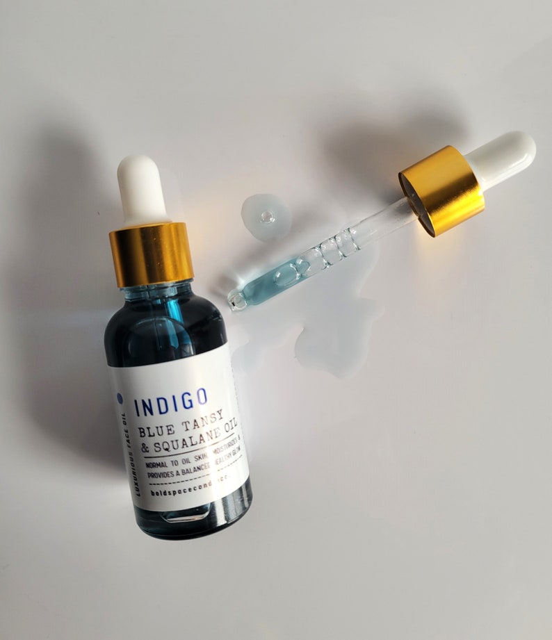 May include: A dark blue glass bottle of Indigo Blue Tansy & Squalane Oil with a white label and gold cap. A glass dropper with a gold top is next to the bottle, with a small puddle of oil. The product is for normal to oily skin.