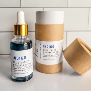May include: A clear glass bottle with a gold dropper and blue liquid, labeled "INDIGO Blue Tansy & Squalane Oil." Beside it, a cylindrical cardboard container with the same label. The product is designed for normal to oily skin.