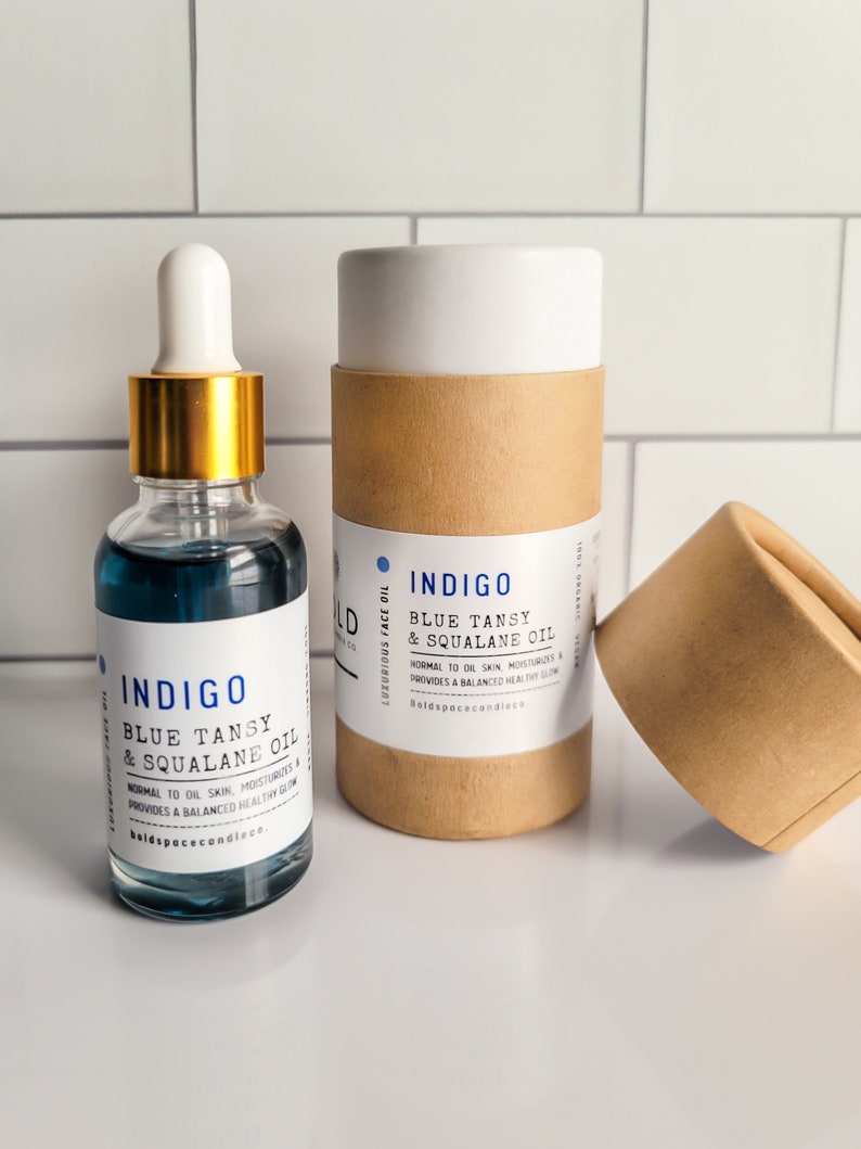 May include: A clear glass bottle with a gold dropper and blue liquid, labeled "INDIGO Blue Tansy & Squalane Oil." Beside it, a cylindrical cardboard container with the same label. The product is designed for normal to oily skin.