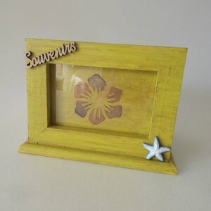 Peut inclure: Un cadre photo en bois jaune avec un devant en verre et une étoile de mer blanche en bas à droite. Le cadre a une inscription en bois découpée qui dit "Souvenirs" en haut à gauche. Le cadre a un motif floral à l'intérieur.