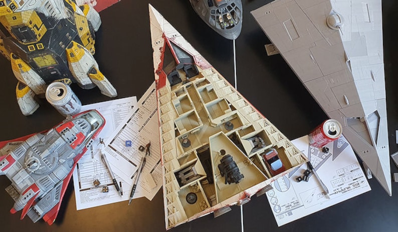 May include: A collection of model spaceships, including a white and red spaceship with an open interior, a gray spaceship, and a yellow and black spaceship. The spaceships are on a table with paper instructions and tools for building models.
