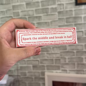 May include: A rectangular white box with red text that reads Spark the middle and break in half. The packaging includes detailed manufacturer information from TABI Cat Curiosities, listing usage instructions, wish limitations, and long-term effects.