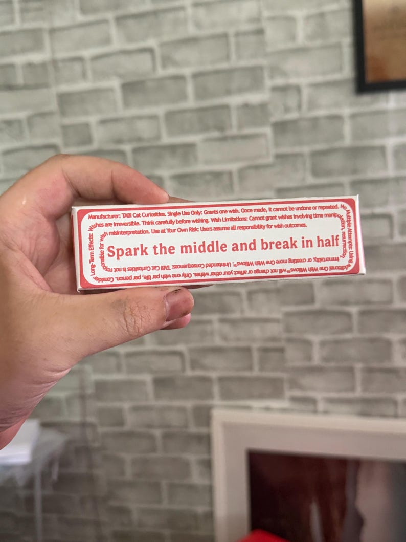 May include: A rectangular white box with red text that reads Spark the middle and break in half. The packaging includes detailed manufacturer information from TABI Cat Curiosities, listing usage instructions, wish limitations, and long-term effects.