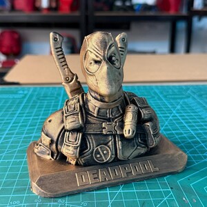 May include: A bronze-colored Deadpool bust, featuring the character's mask and costume details. The bust is mounted on a rectangular base with the word "DEADPOOL" engraved on it. The figure has two swords on its back.