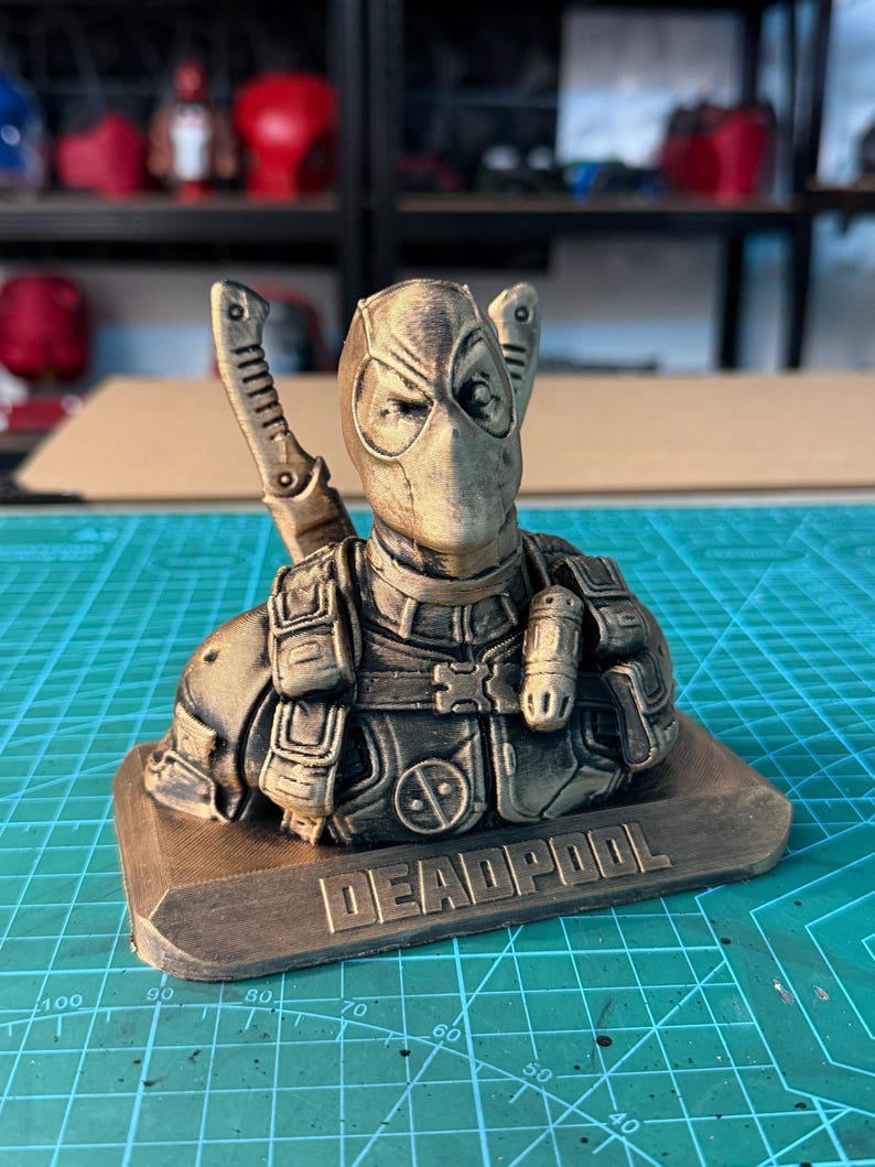 May include: A bronze-colored Deadpool bust, featuring the character's mask and costume details. The bust is mounted on a rectangular base with the word "DEADPOOL" engraved on it. The figure has two swords on its back.