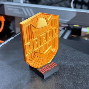 May include: Orange 3D-printed shield with the word "DREDD" in bold letters. The shield is mounted on a black rectangular base with the word "DREDD" in red. The design features geometric shapes and lines, suggesting a badge or emblem.