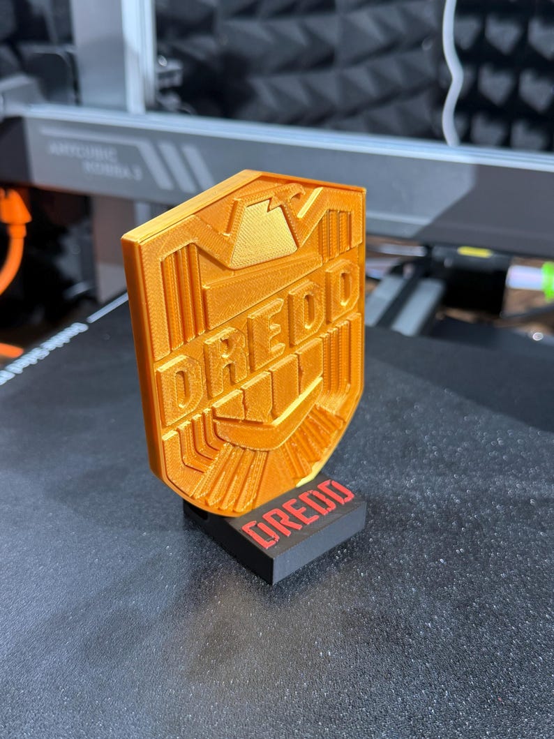 May include: Orange 3D-printed shield with the word "DREDD" in bold letters. The shield is mounted on a black rectangular base with the word "DREDD" in red. The design features geometric shapes and lines, suggesting a badge or emblem.