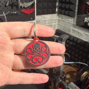 K&ouml;nnte beinhalten: Ein runder Schl&uuml;sselanh&auml;nger mit einem roten und schwarzen Hydra-Symbol, das einen Sch&auml;del und ein Oktopus-Design zeigt. Er ist an einem silbernen Ring befestigt. Der Hintergrund umfasst weitere dekorative Gegenst&auml;nde.