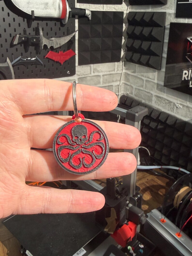 K&ouml;nnte beinhalten: Ein runder Schl&uuml;sselanh&auml;nger mit einem roten und schwarzen Hydra-Symbol, das einen Sch&auml;del und ein Oktopus-Design zeigt. Er ist an einem silbernen Ring befestigt. Der Hintergrund umfasst weitere dekorative Gegenst&auml;nde.
