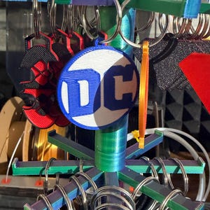 K&ouml;nnte beinhalten: Ein mehrfarbiger Ausstellungsst&auml;nder mit Schl&uuml;sselanh&auml;ngern. Die Schl&uuml;sselanh&auml;nger umfassen ein DC-Logo, ein rot-schwarzes Spinnen-Design und andere Designs. Der St&auml;nder hat blaue, gr&uuml;ne und violette Akzente. Die Schl&uuml;sselanh&auml;nger sind an silbernen Metallringen befestigt.