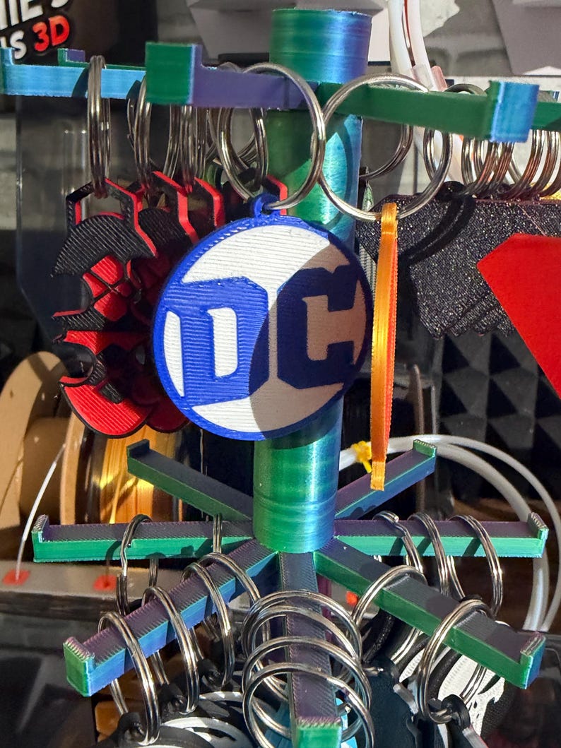 K&ouml;nnte beinhalten: Ein mehrfarbiger Ausstellungsst&auml;nder mit Schl&uuml;sselanh&auml;ngern. Die Schl&uuml;sselanh&auml;nger umfassen ein DC-Logo, ein rot-schwarzes Spinnen-Design und andere Designs. Der St&auml;nder hat blaue, gr&uuml;ne und violette Akzente. Die Schl&uuml;sselanh&auml;nger sind an silbernen Metallringen befestigt.
