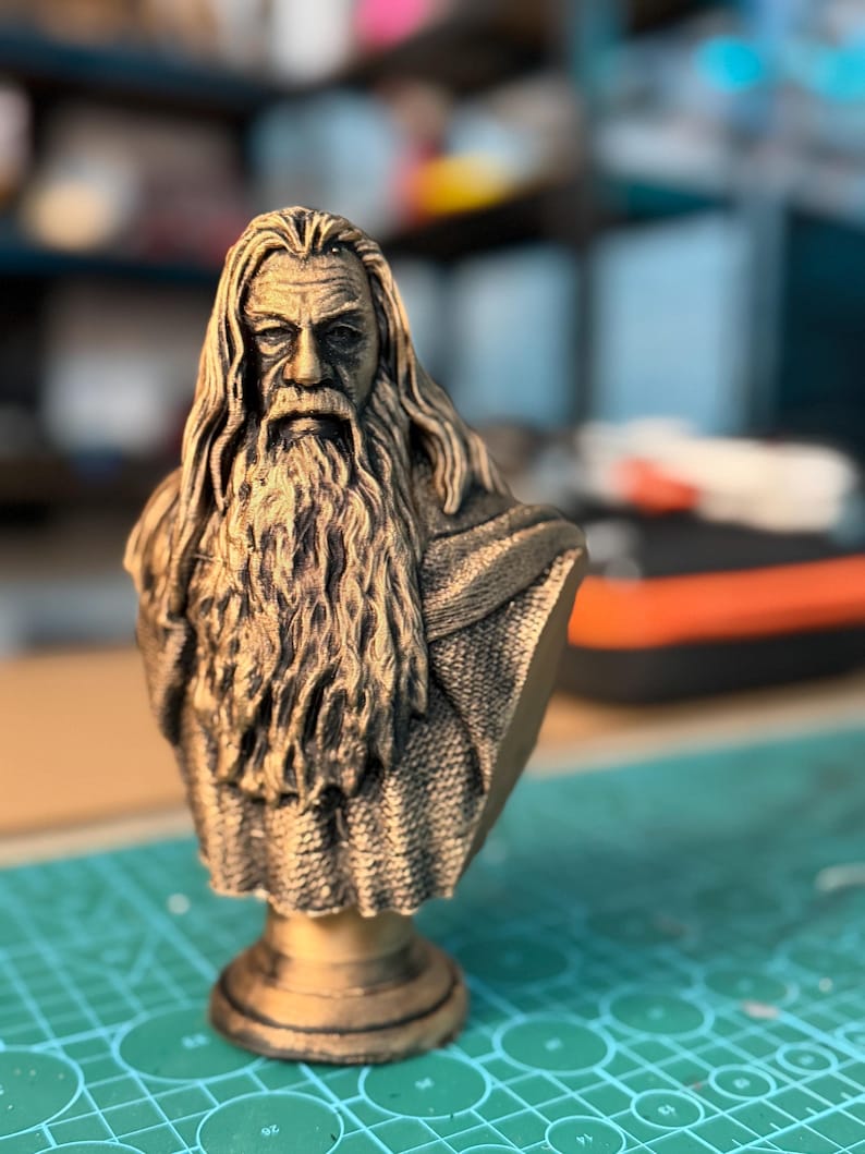 May include: A bronze-colored bust of a wise, elderly man with a long beard and flowing hair. The sculpture is detailed, with visible wrinkles and textures. The bust is mounted on a pedestal, and the background is blurred.