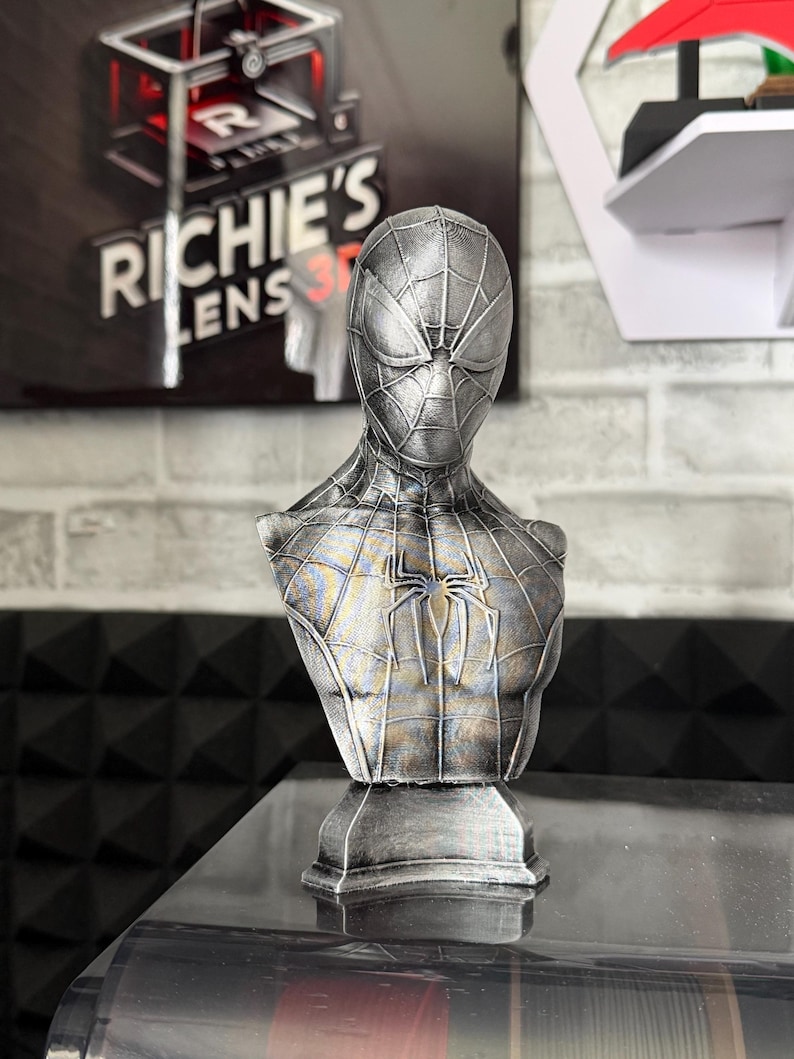 May include: A silver-colored Spider-Man bust, featuring the character's mask and torso. The bust has a textured finish and a spider emblem on the chest. The bust is displayed on a black surface.