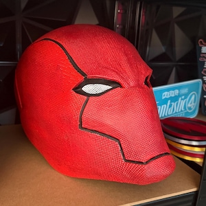 Op de afbeelding: Een felrode helm met zwarte details en een wit gaas ooggedeelte. De helm wordt op een plank tentoongesteld. Het ontwerp doet denken aan een stripboekpersonage.