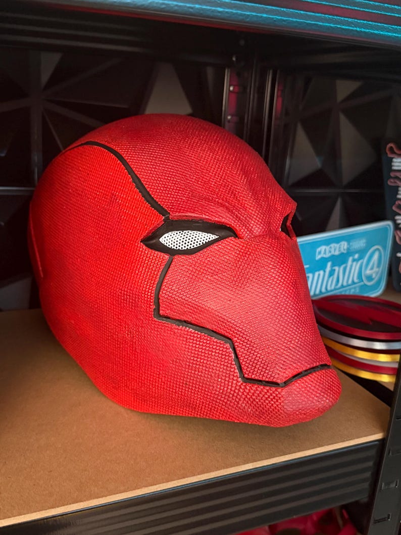 Op de afbeelding: Een felrode helm met zwarte details en een wit gaas ooggedeelte. De helm wordt op een plank tentoongesteld. Het ontwerp doet denken aan een stripboekpersonage.