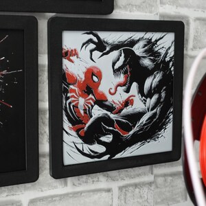 May include: A square black-framed wall art piece featuring a high-contrast graphic illustration of Spider-Man in red and black facing off against the character Venom in black and white with sharp claws and a long tongue, set against a white background.