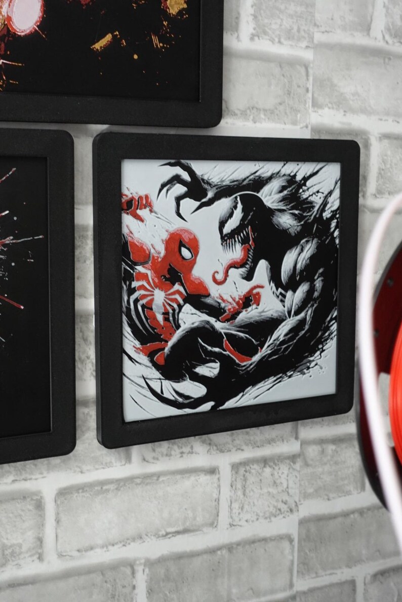 May include: A square black-framed wall art piece featuring a high-contrast graphic illustration of Spider-Man in red and black facing off against the character Venom in black and white with sharp claws and a long tongue, set against a white background.