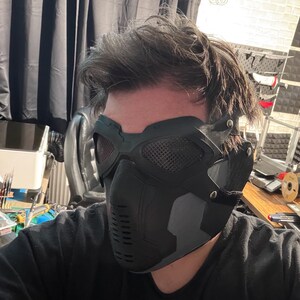 May include: A black and gray tactical mask with mesh eye coverings and a contoured design. The mask features a series of vents and a secure strap system, offering a futuristic aesthetic.