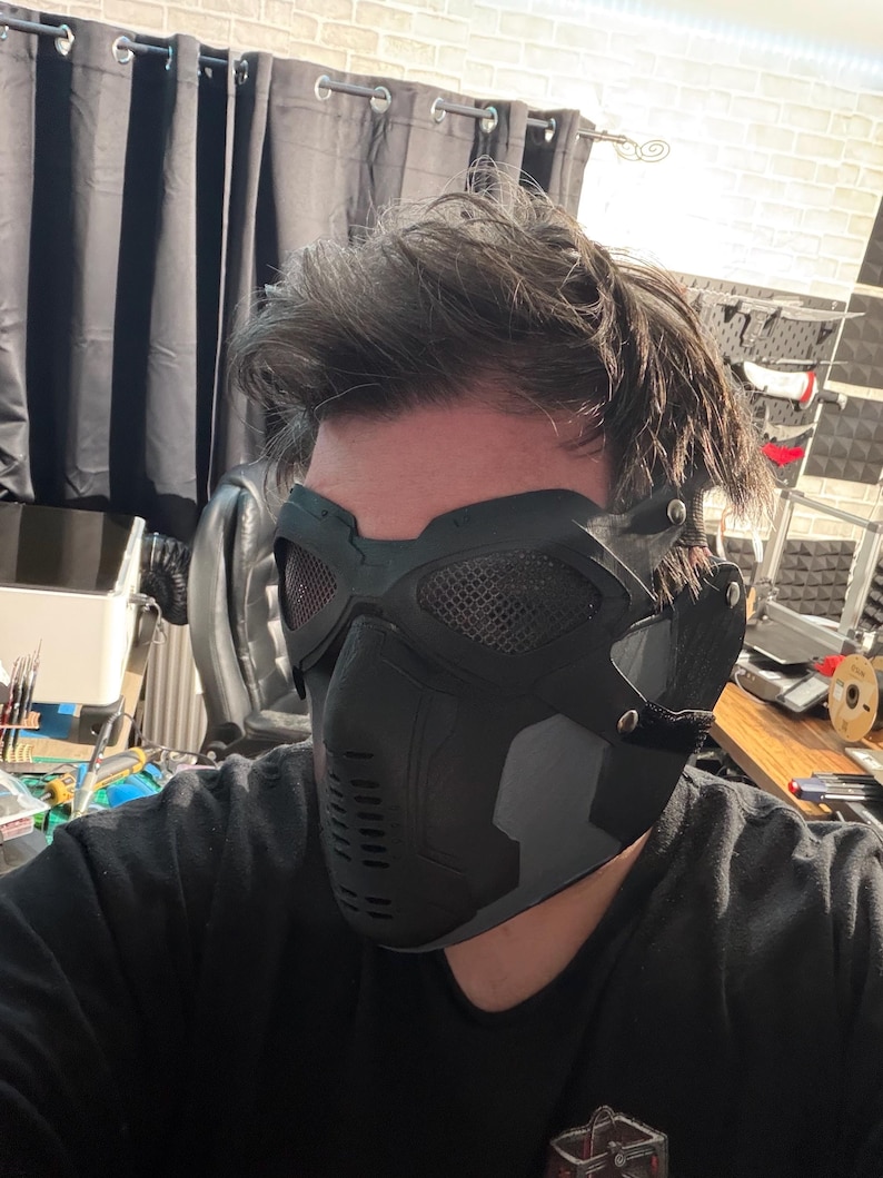 May include: A black and gray tactical mask with mesh eye coverings and a contoured design. The mask features a series of vents and a secure strap system, offering a futuristic aesthetic.
