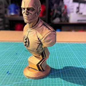 May include: A bronze-colored bust of The Flash superhero. The bust features detailed facial features, a mask, and the Flash emblem on the chest. The sculpture is mounted on a geometric base, showcasing a textured surface.