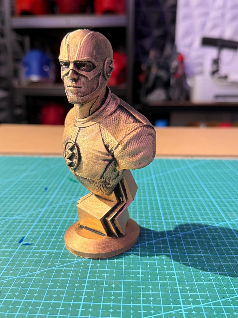 May include: A bronze-colored bust of The Flash superhero. The bust features detailed facial features, a mask, and the Flash emblem on the chest. The sculpture is mounted on a geometric base, showcasing a textured surface.
