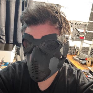 May include: Black tactical mask with mesh eye coverings and a vented lower face section. The mask is made of a matte black material and has a strap for securing it around the head. The design is reminiscent of a futuristic or military aesthetic.