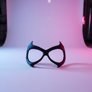 May include: A black eye mask with a smooth, matte finish, featuring two large, rounded eye openings and pointed, upward-curving edges at the top and bottom, designed to cover the eye area with a distinct, symmetrical, and angular silhouette.
