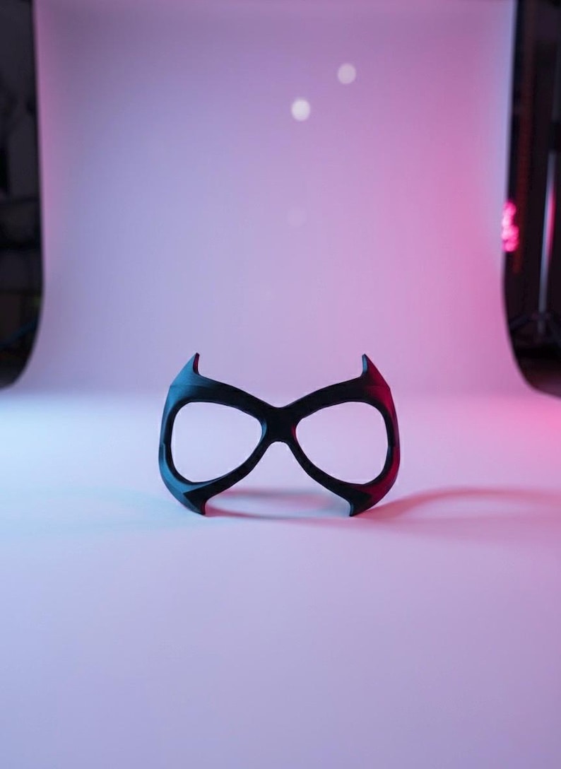 May include: A black eye mask with a smooth, matte finish, featuring two large, rounded eye openings and pointed, upward-curving edges at the top and bottom, designed to cover the eye area with a distinct, symmetrical, and angular silhouette.
