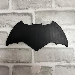 May include: A black, flat, bat-shaped emblem featuring a pointed top and curved wings. The surface of the object displays a fine, textured grid pattern consistent with 3D printing, creating a matte finish across the entire silhouette of the bat.