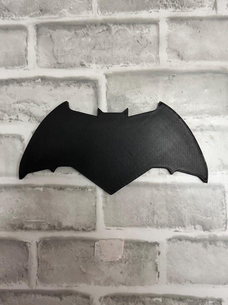 May include: A black, flat, bat-shaped emblem featuring a pointed top and curved wings. The surface of the object displays a fine, textured grid pattern consistent with 3D printing, creating a matte finish across the entire silhouette of the bat.