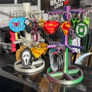 May include: A multi-tiered display stand holding a collection of character keyrings. Keyrings feature designs such as Spider-Man, Ghostface, and Superman. The stand is in various colours, including red, blue, and green, and is designed to hold multiple keyrings.