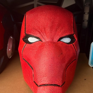 Op de afbeelding: Een close-up van een rode, getextureerde masker met zwarte randen en witte mesh oogopeningen. Het masker is een replica van een superheldenhelm, waarschijnlijk voor cosplay of display.