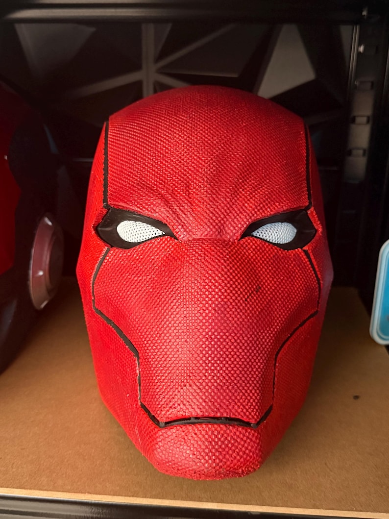 Op de afbeelding: Een close-up van een rode, getextureerde masker met zwarte randen en witte mesh oogopeningen. Het masker is een replica van een superheldenhelm, waarschijnlijk voor cosplay of display.