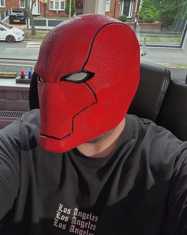 Op de afbeelding: Een felrood masker met een getextureerd oppervlak en zwarte contouren. Het masker wordt gedragen met een zwart t-shirt met de tekst "Los Angeles" in een gestileerd lettertype.