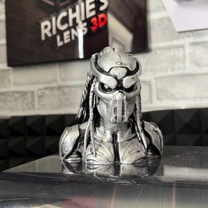 May include: A silver-colored Predator bust, a detailed 3D-printed sculpture. The bust features the character's iconic mask, dreadlocks, and armor. The background includes a sign that reads "RICHIE'S LENS 3D".